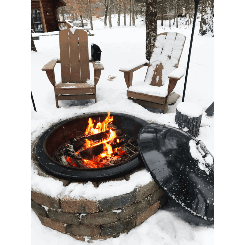 A fire burns in the Legacy Fire Pit Insert surrounded by snow-covered chairs and ground. The Snuffer Lid protects the inside of the fire pit from moisture to ensure that the dry fire pit is ready for the next fire in any weather.