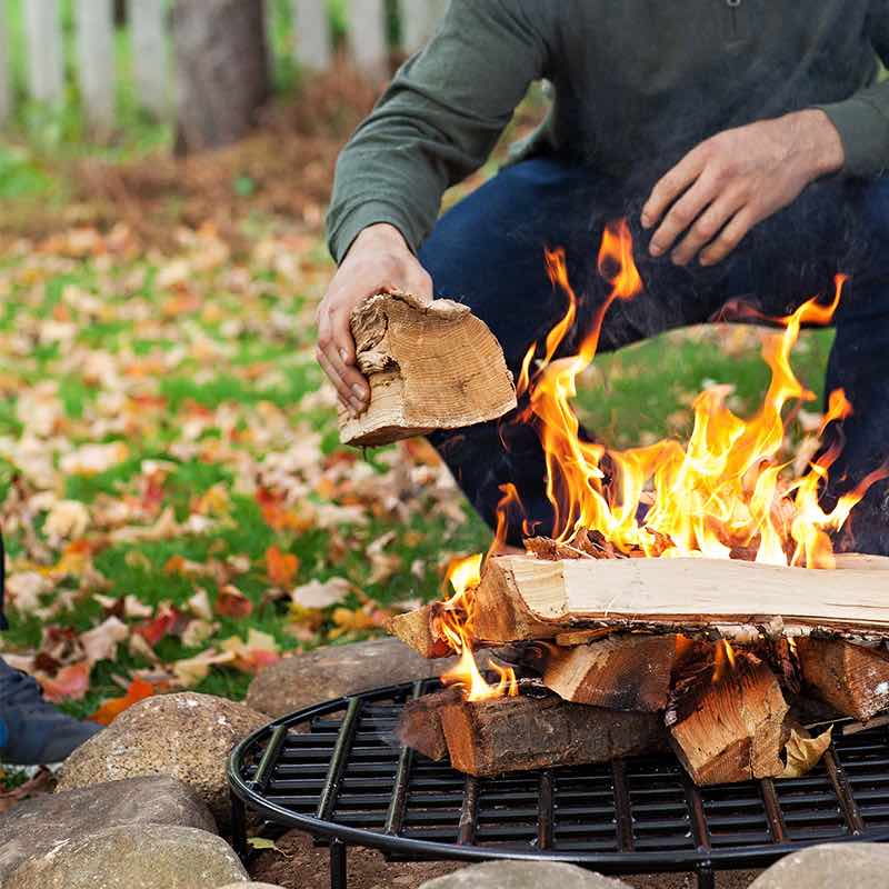 A man feeding split logs onto a fire built on a Walden Original Fire Grate, autumn leaves on the lawn behind
