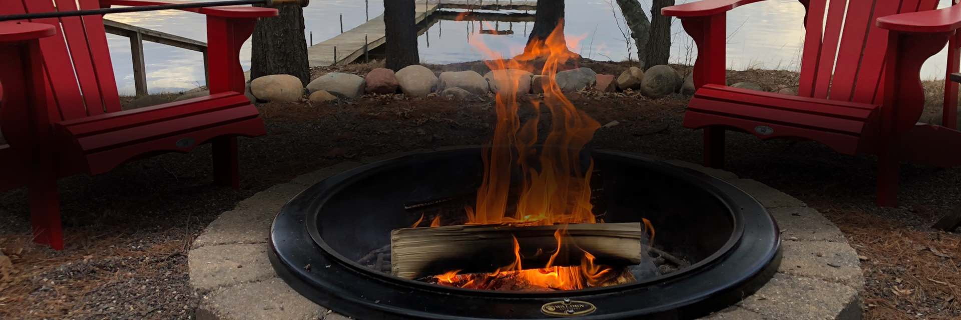 Roaring fire inside a lakeside fire pit with two lounge chairs behind.