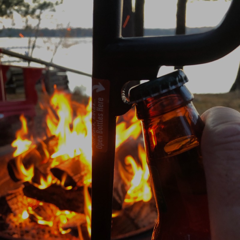 Bottle of beer being opened with a bottle opener built into a fire poker with bonfire in the background