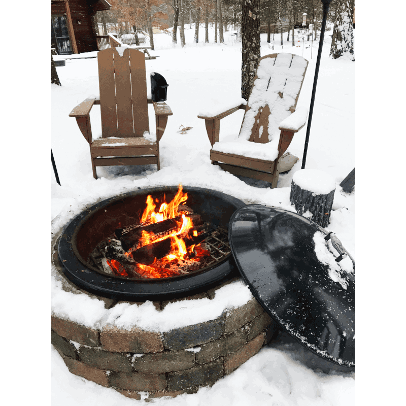 A fire burns in the Legacy Fire Pit Insert surrounded by snow-covered chairs and ground. The Snuffer Lid protects the inside of the fire pit from moisture to ensure that the dry fire pit is ready for the next fire in any weather.