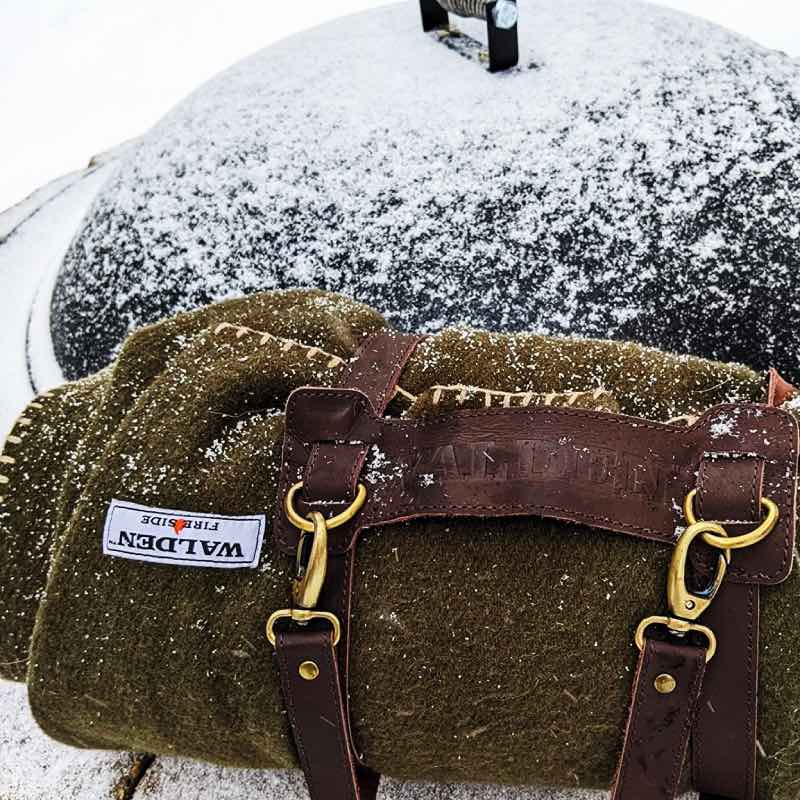 Fire retardant Walden wool blanket rolled up and held by its leather strap, laying on top of an outdoor fire pit covered with a snow dusted lid.